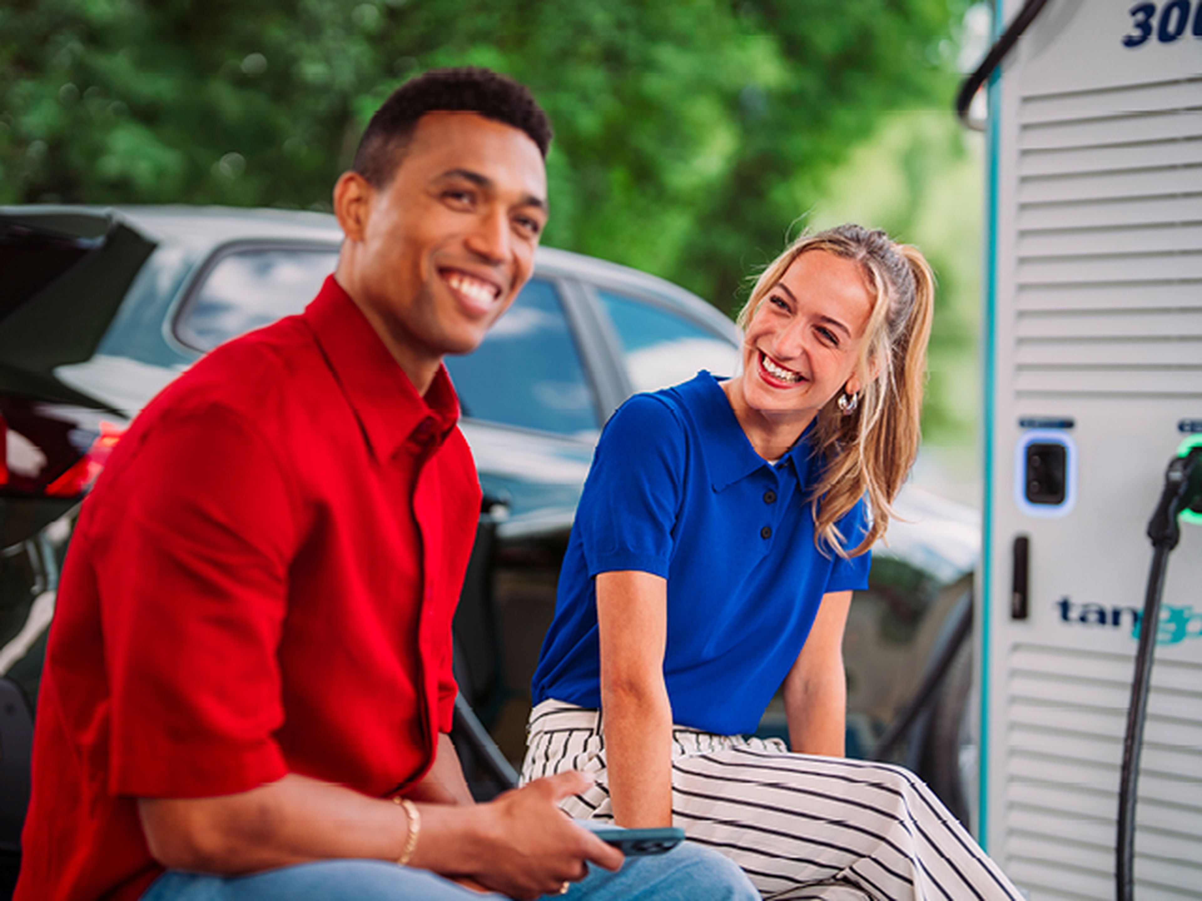 Jonge man en vrouw zitten en glimlachen op een bank tijden het laden van hun auto bij Tango electric