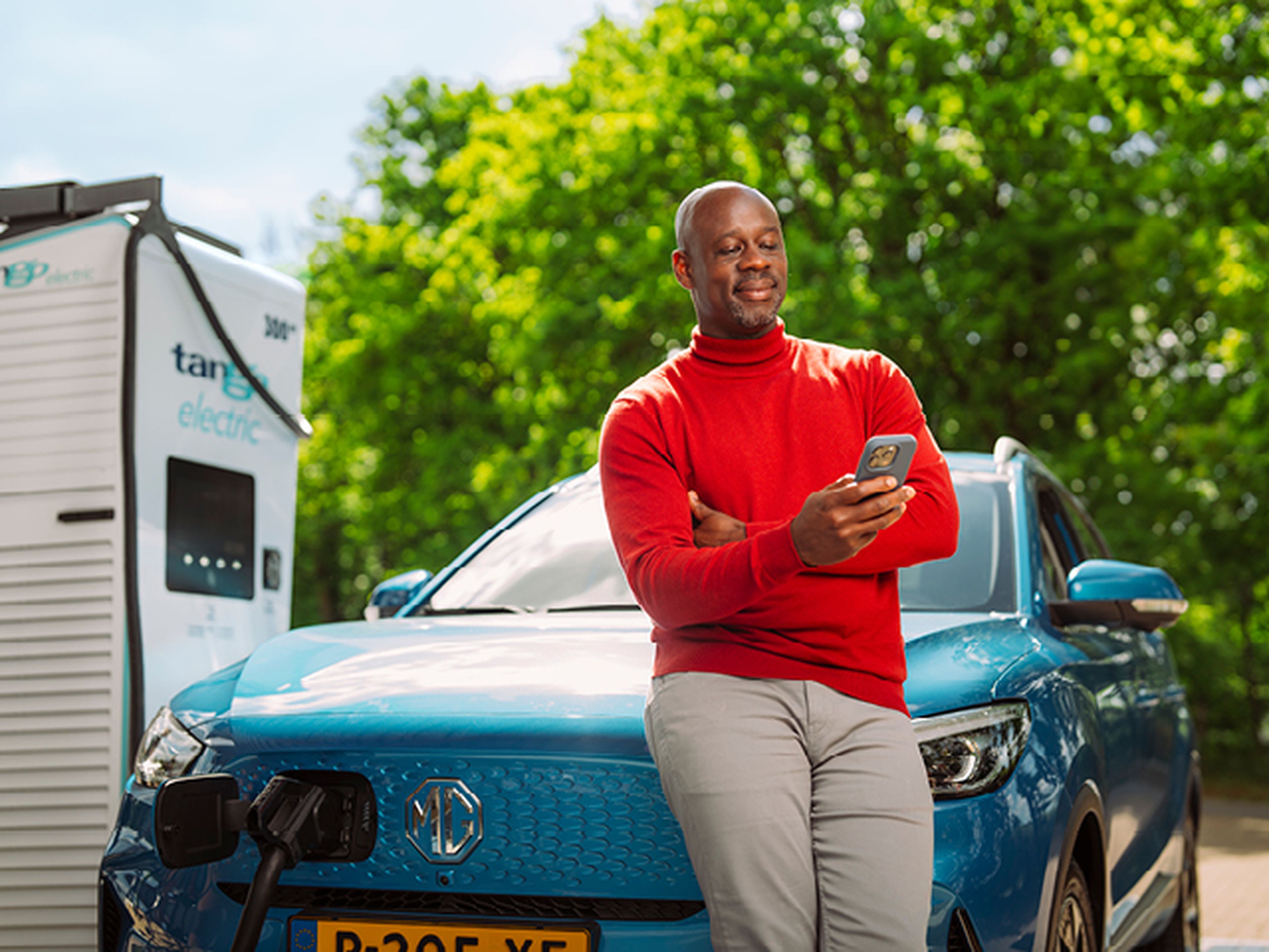 Een man in een rode trui kijkt op zijn smartphone en leunt ontspannen tegen een blauwe elektrische auto terwijl deze oplaadt bij een Tango electric laadstation.