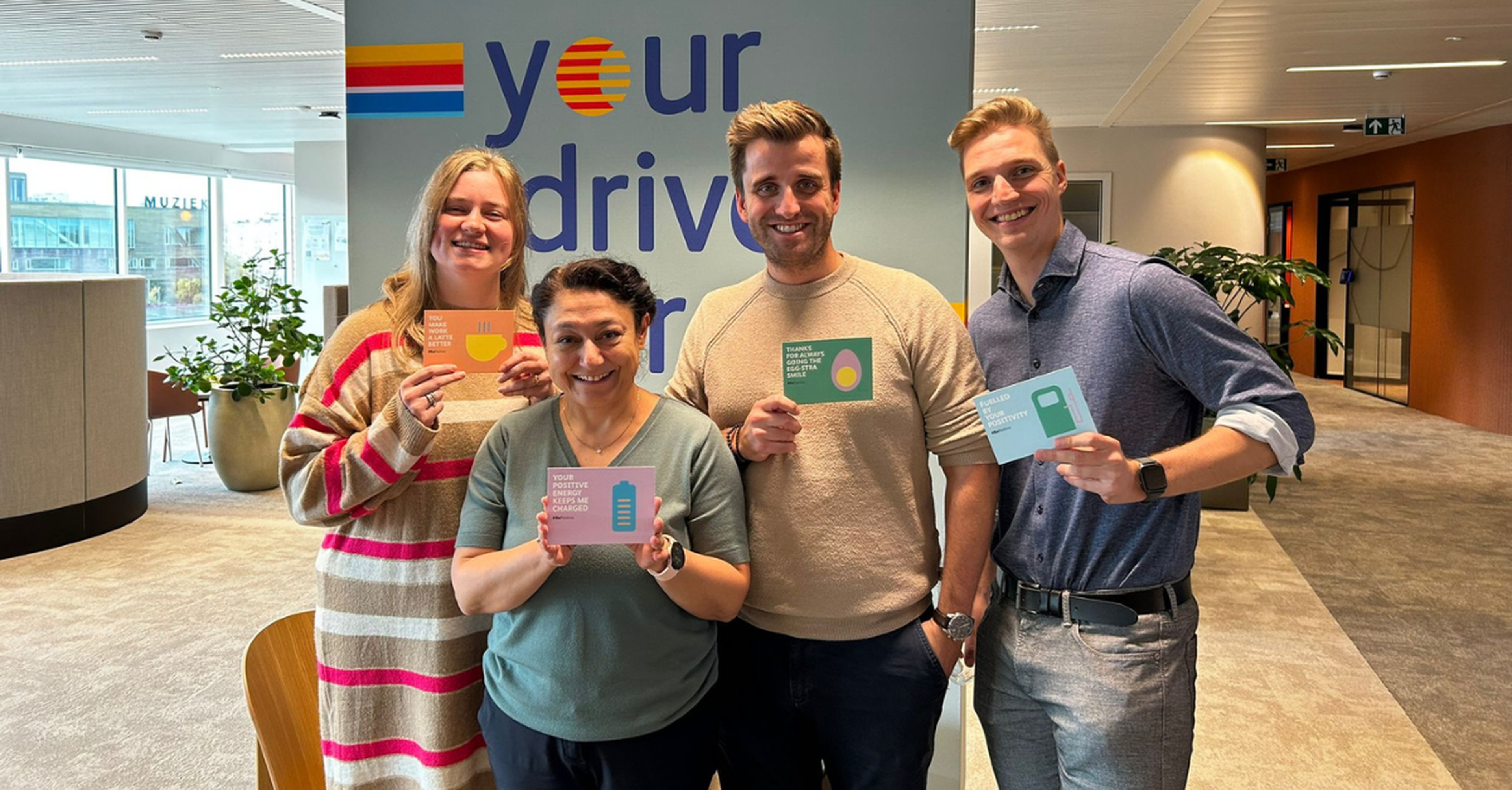 Een groep van 4 collega's glimlacht naar de camera terwijl ze een #BePositive kaartje vasthouden.