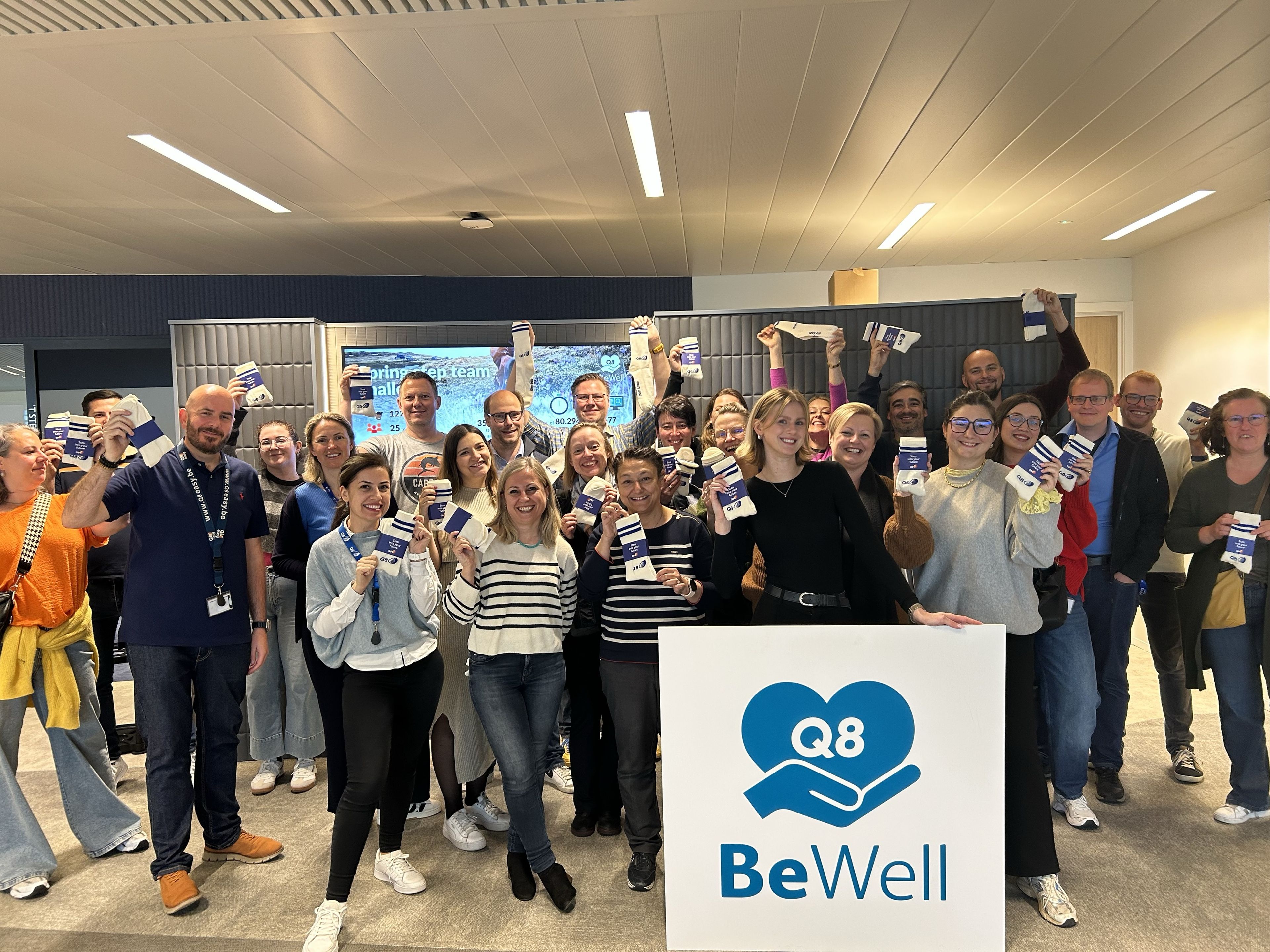 Een grote groep Q8 collega's glimlacht voor de camera en neemt een groepsfoto met een BeWell bord ervoor.