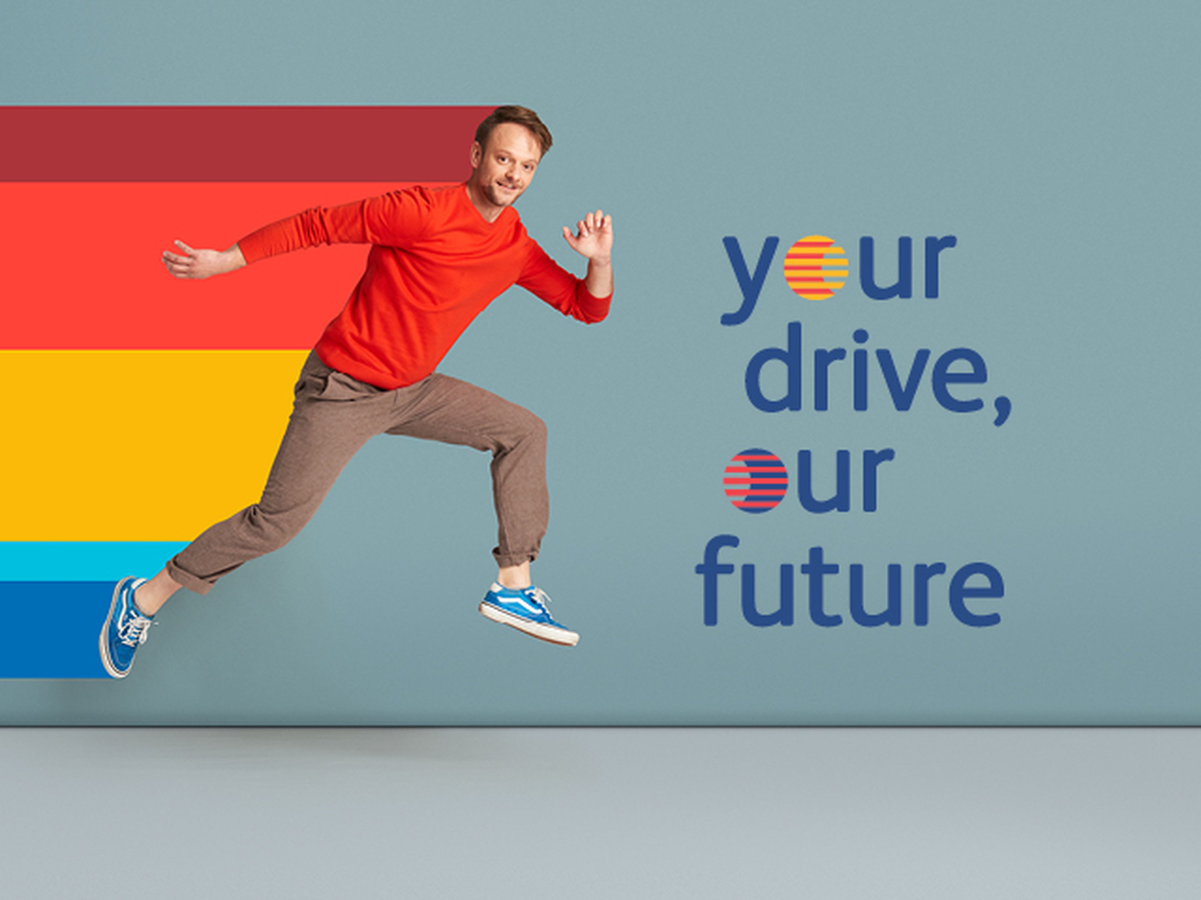 Een persoon springt energiek in de lucht tegen een effen blauwe achtergrond en draagt een felrode trui, opgerolde bruine broek en blauwe sneakers.