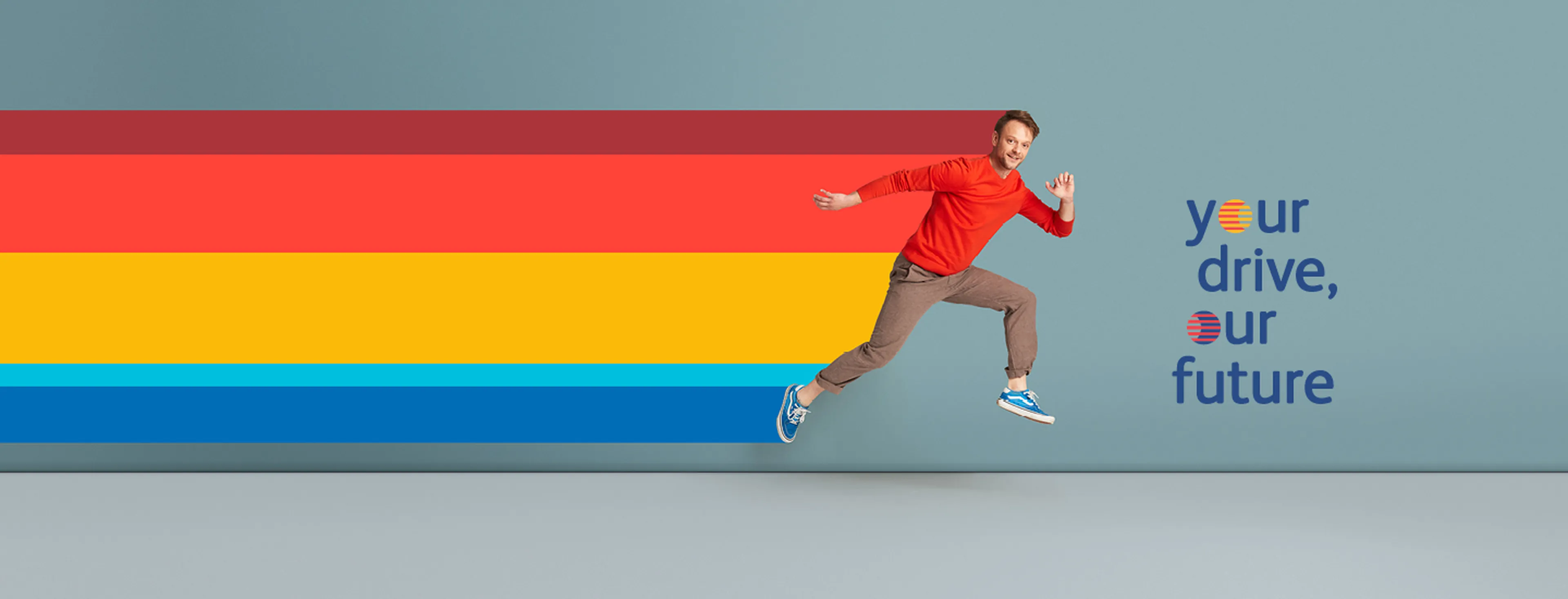 Een persoon springt energiek in de lucht tegen een effen blauwe achtergrond en draagt een felrode trui, opgerolde bruine broek en blauwe sneakers.