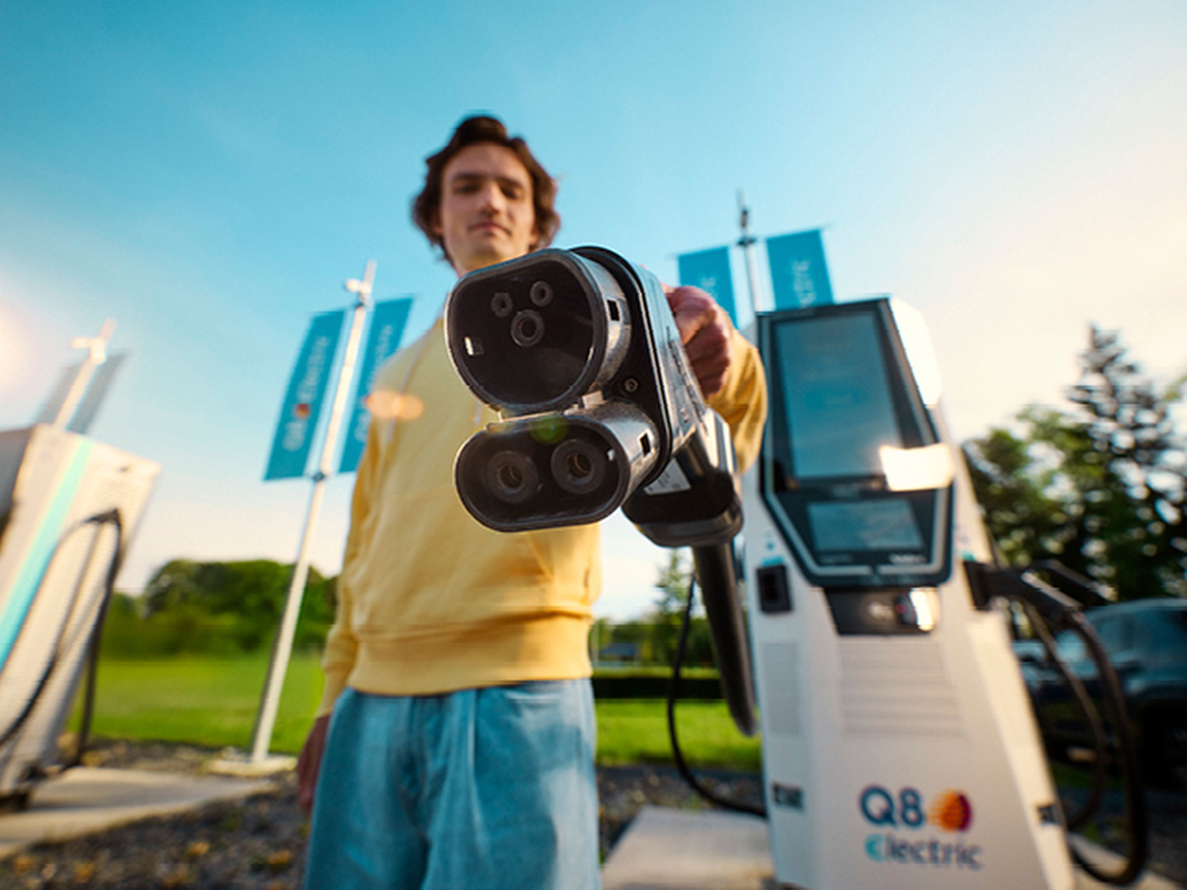 Een persoon in een gele trui houdt een EV-laadstekker naar de camera bij een Q8 electric-laadstation.
