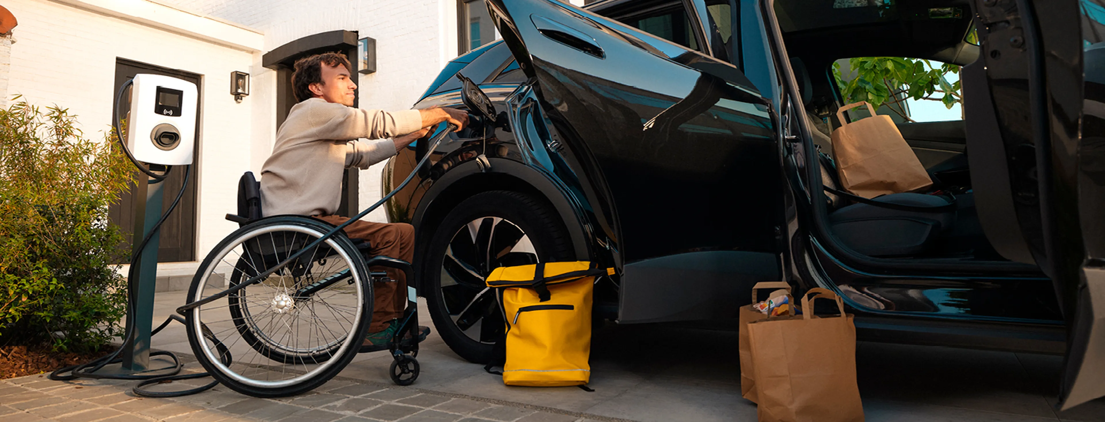 Een man in een rolstoel sluit zijn elektrische auto aan via een thuislader bij zijn woning.