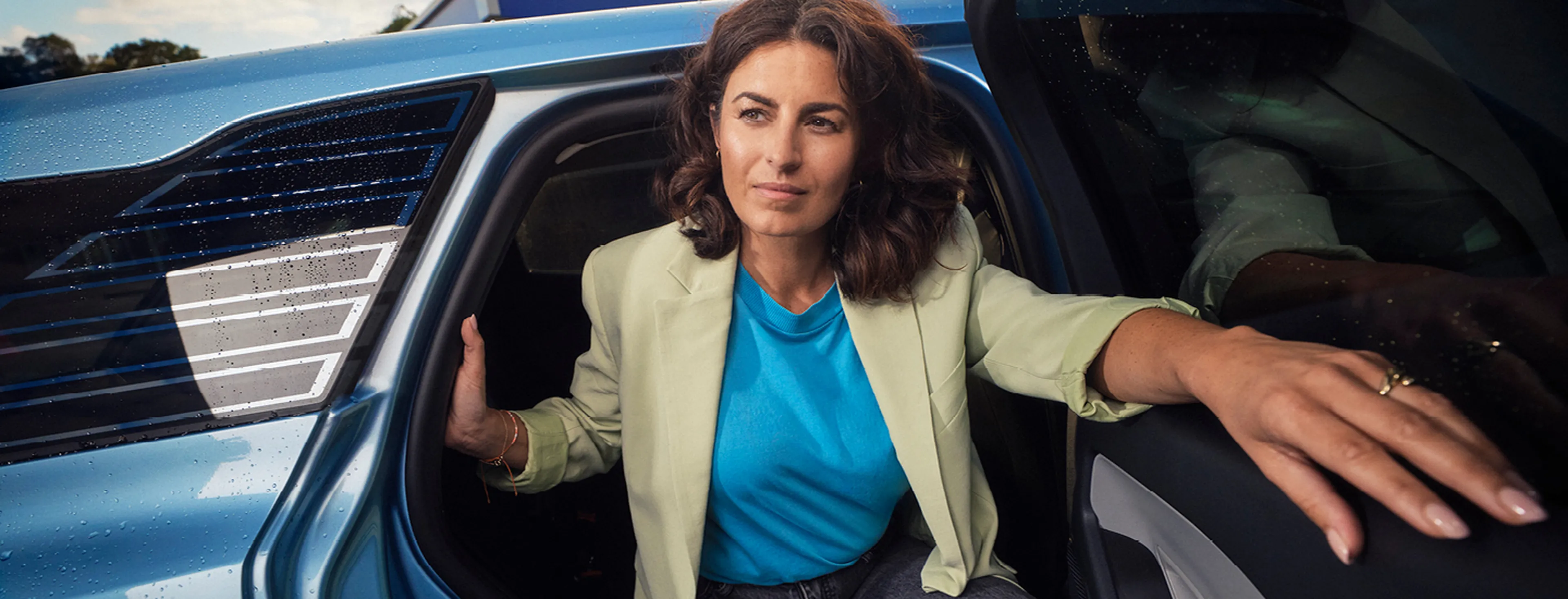 Een vrouw met donker krullend haar stapt uit een blauwe elektrische auto. Ze draagt een beige blazer, een felblauw T-shirt en een donkere jeans.