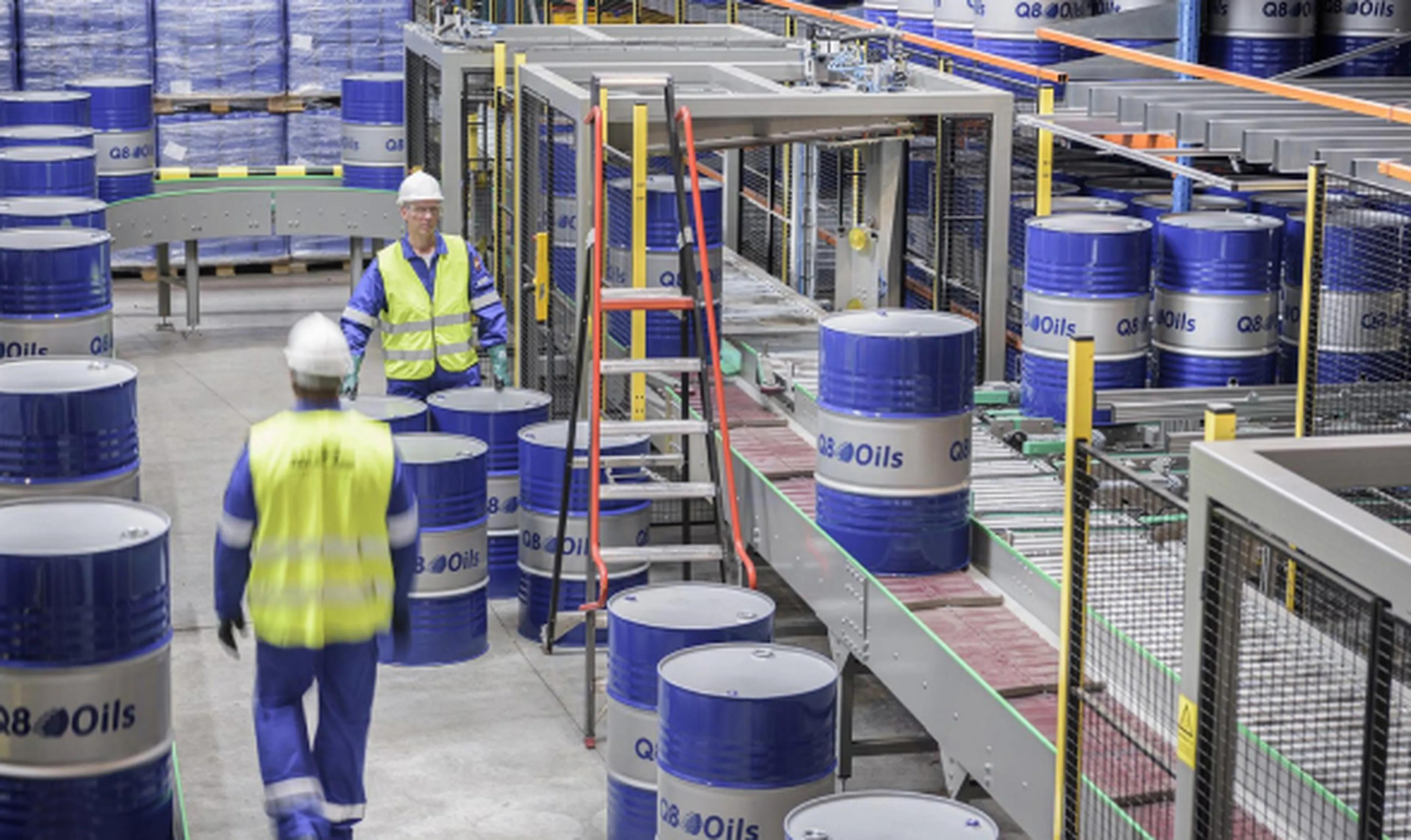 Binnenzicht van de Q8Oils blending plant waar blauwe Q8Oils-vaten op een transportband staan. Twee medewerkers in blauwe overalls, gele veiligheidshesjes en witte helmen lopen tussen de vaten.