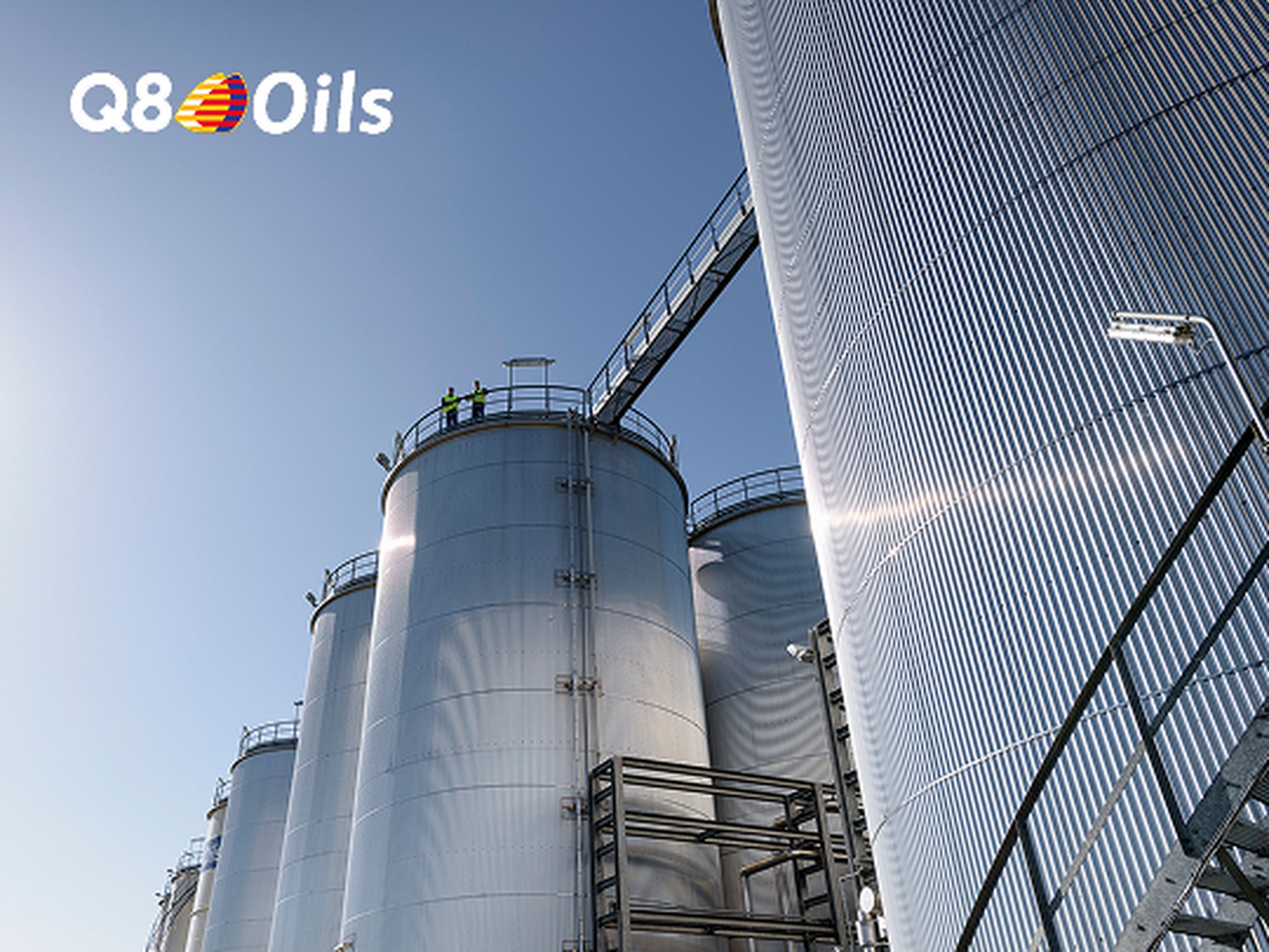 Opslagtanks van een Q8 Oils blending plant torenen op tegen een heldere blauwe lucht. Twee medewerkers in fluohesjes staan boven op een van de tanks.