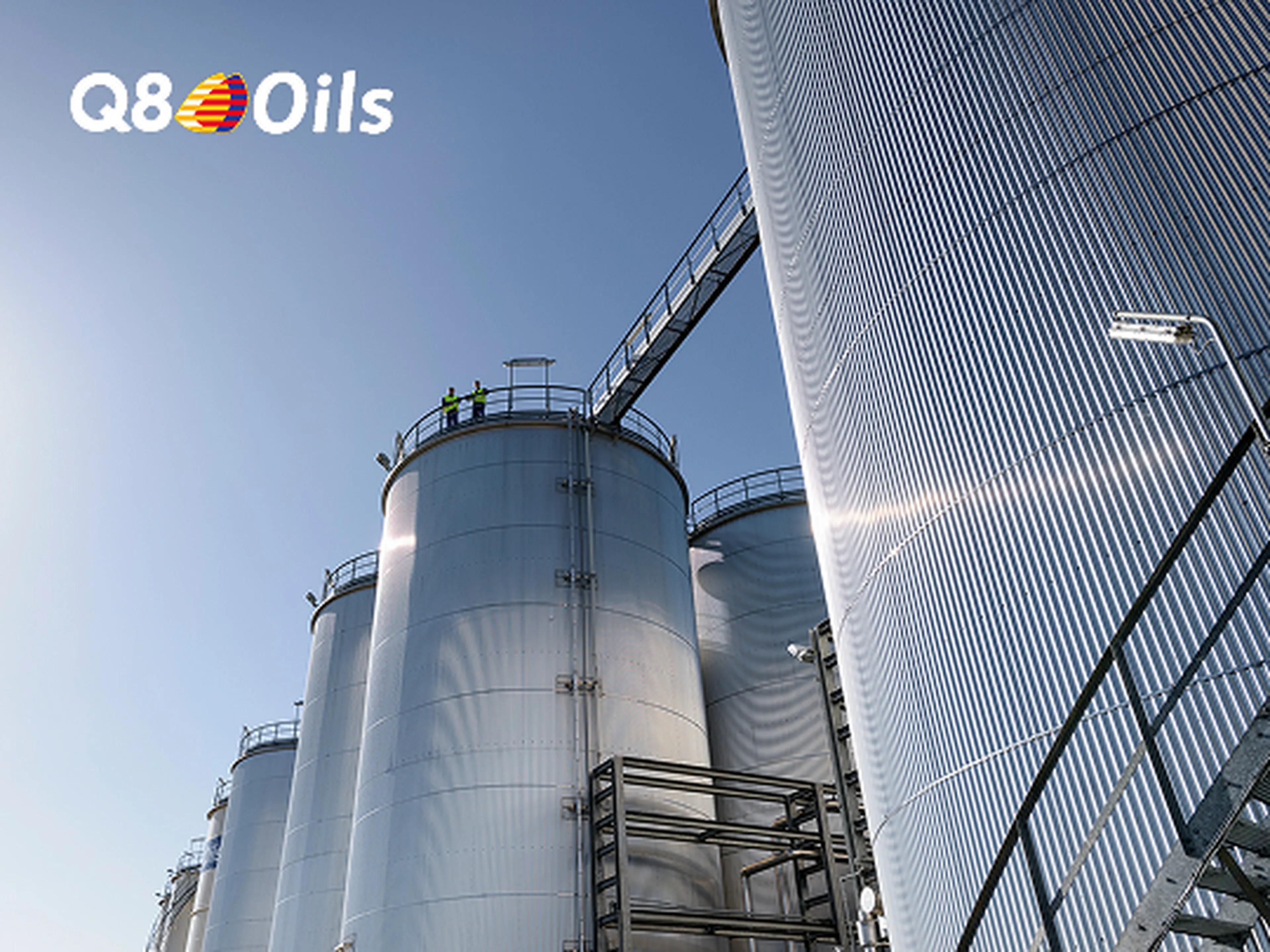 Opslagtanks van een Q8 Oils blending plant torenen op tegen een heldere blauwe lucht. Twee medewerkers in fluohesjes staan boven op een van de tanks.
