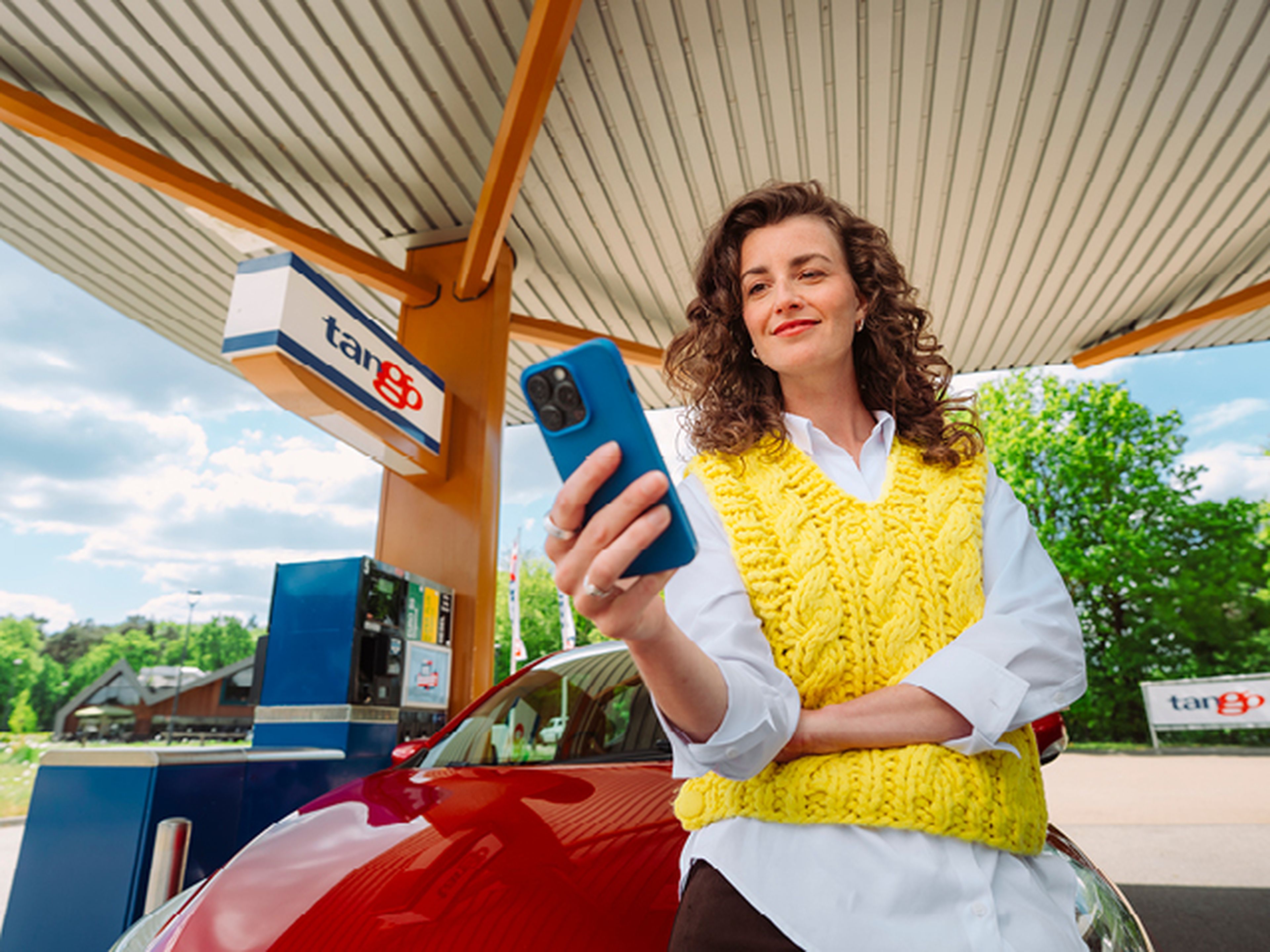Vrouw leunt tegen haar rode auto in Tango tankstation en glimlacht naar haar smartphone