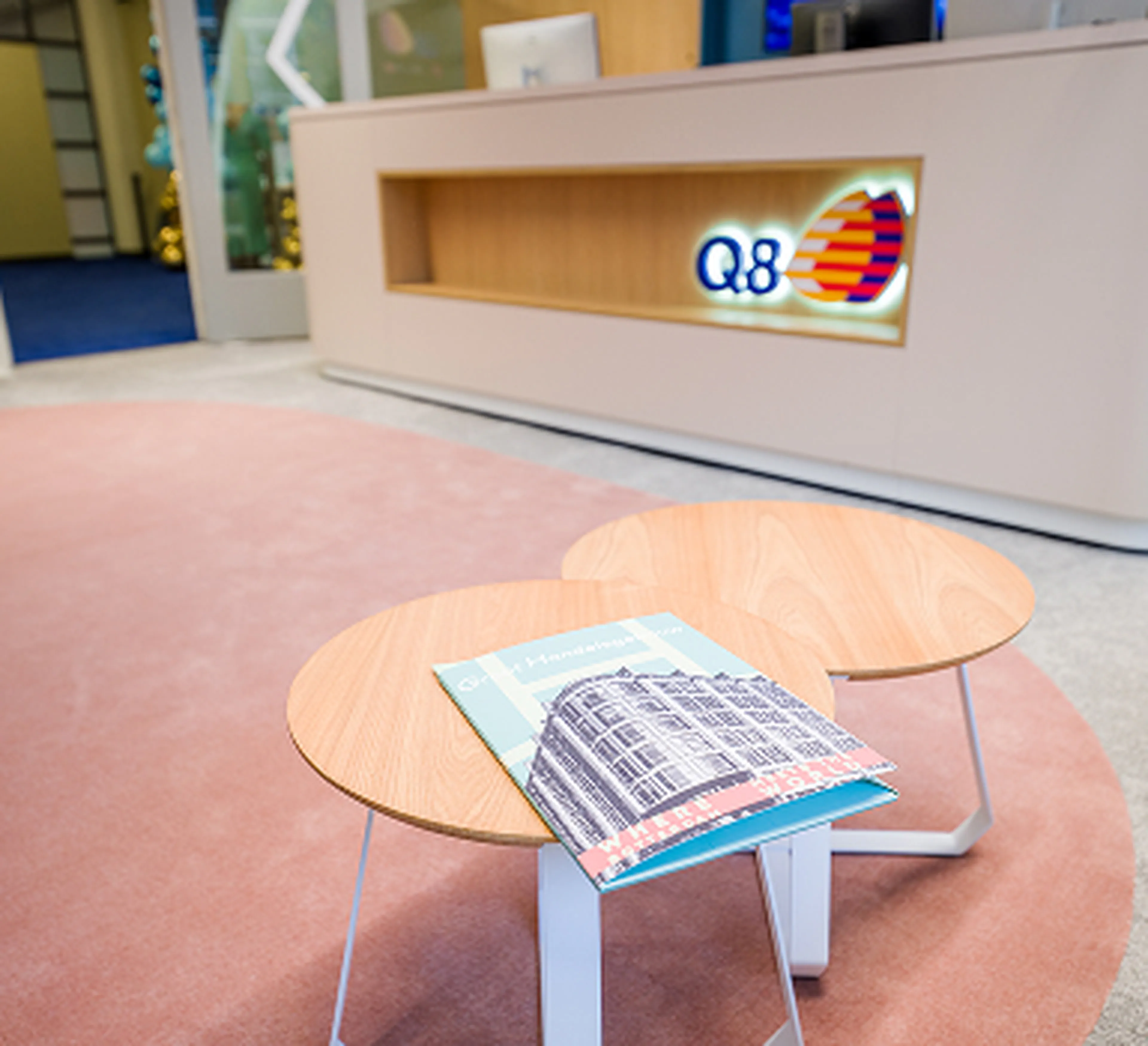 Receptiebalie van Q8 op het hoofdkantoor in Rotterdam met het logo zichtbaar op de wand. Op de voorgrond staan twee ronde houten tafeltjes met een boek erop, op een zachtroze tapijt.