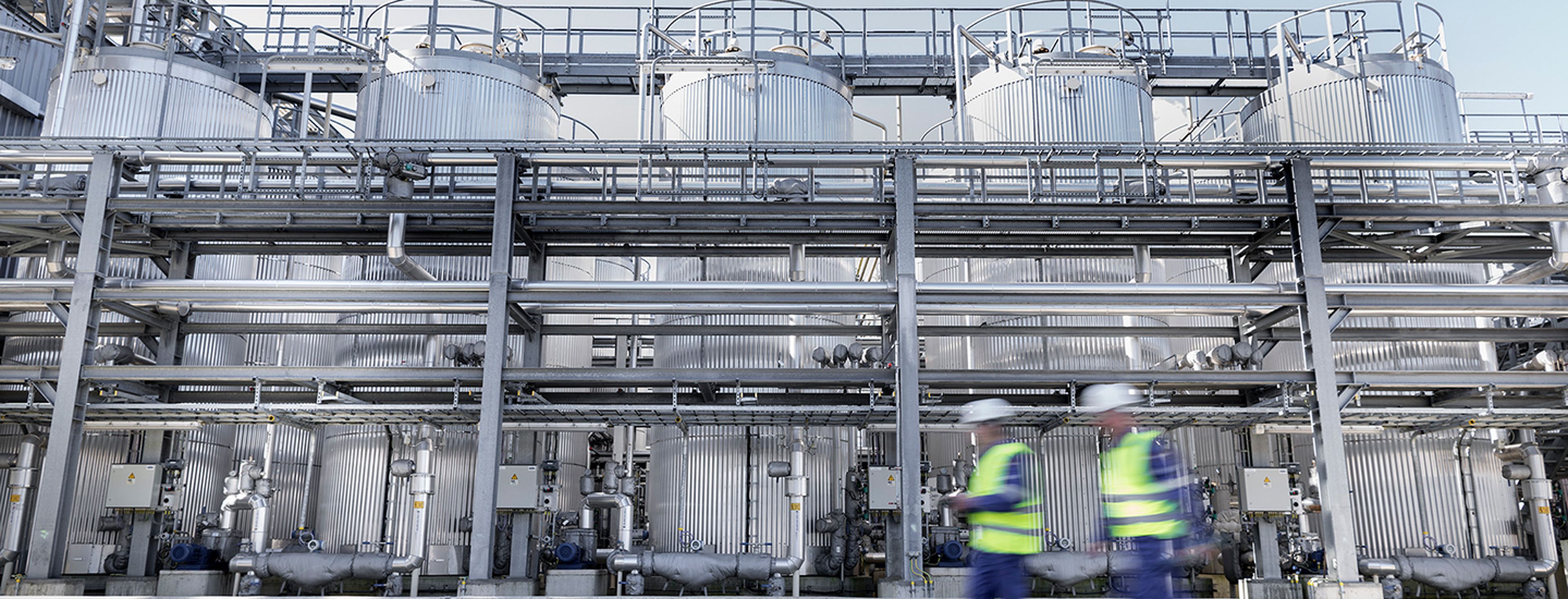 Twee medewerkers in veiligheidskledij wandelen langs een indrukwekkende installatie van verticale opslagtanks en buizen in de Q8 Oils blending plant in Antwerpen.