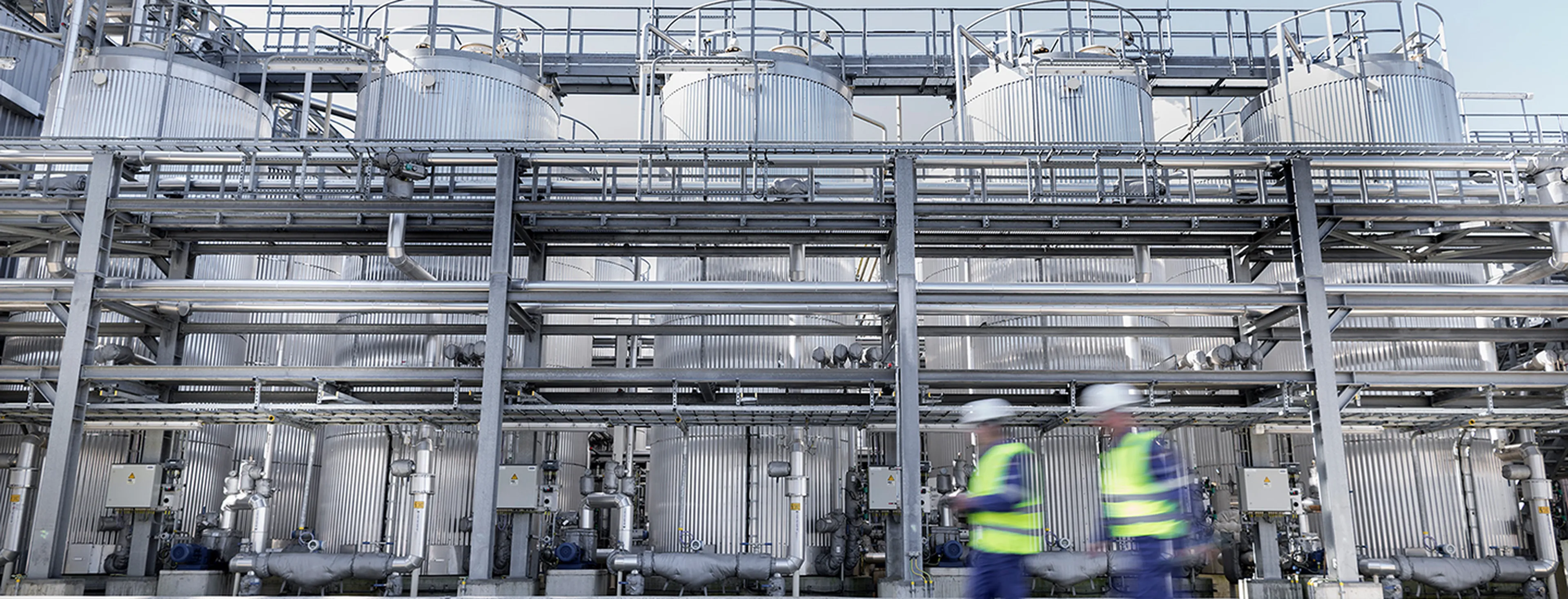 Twee medewerkers in veiligheidskledij wandelen langs een indrukwekkende installatie van verticale opslagtanks en buizen in de Q8 Oils blending plant in Antwerpen.