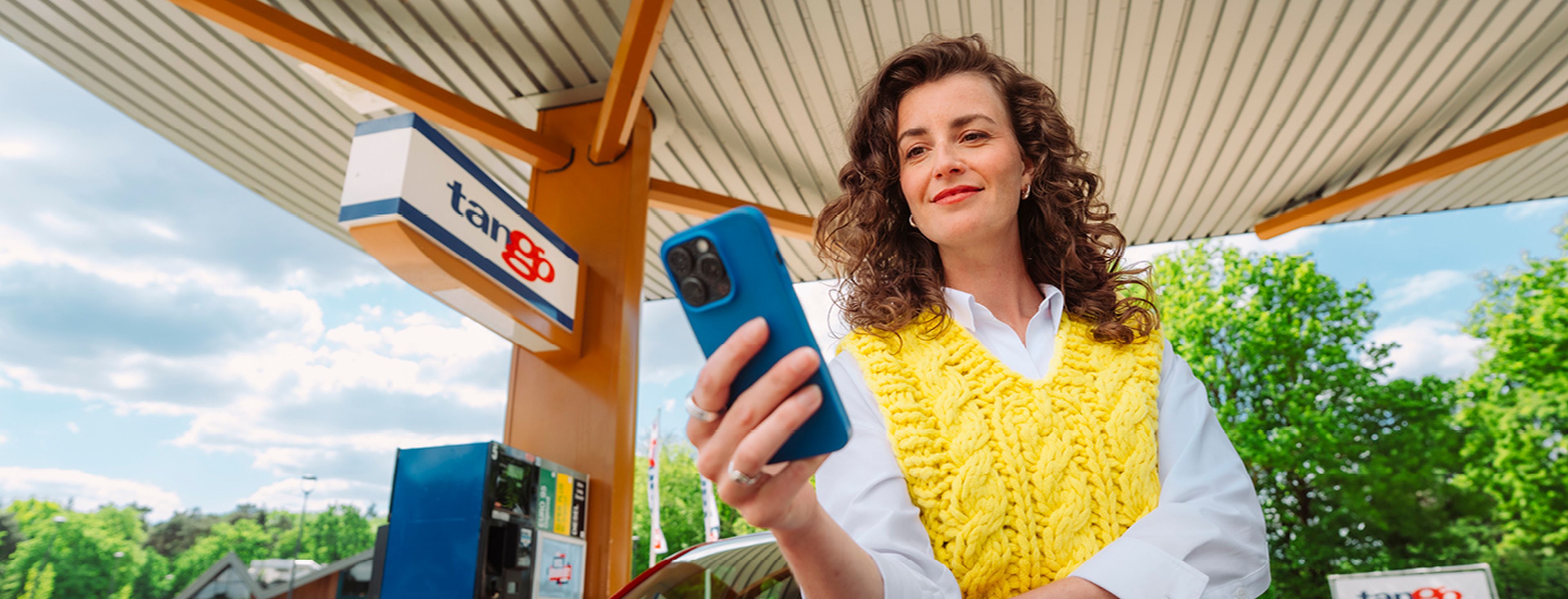 Vrouw bekijkt de Q8 app op hVrouw staat glimlachend bij een Tango tankstation naast haar autoaar telefoon bij een Q8 tankstation.