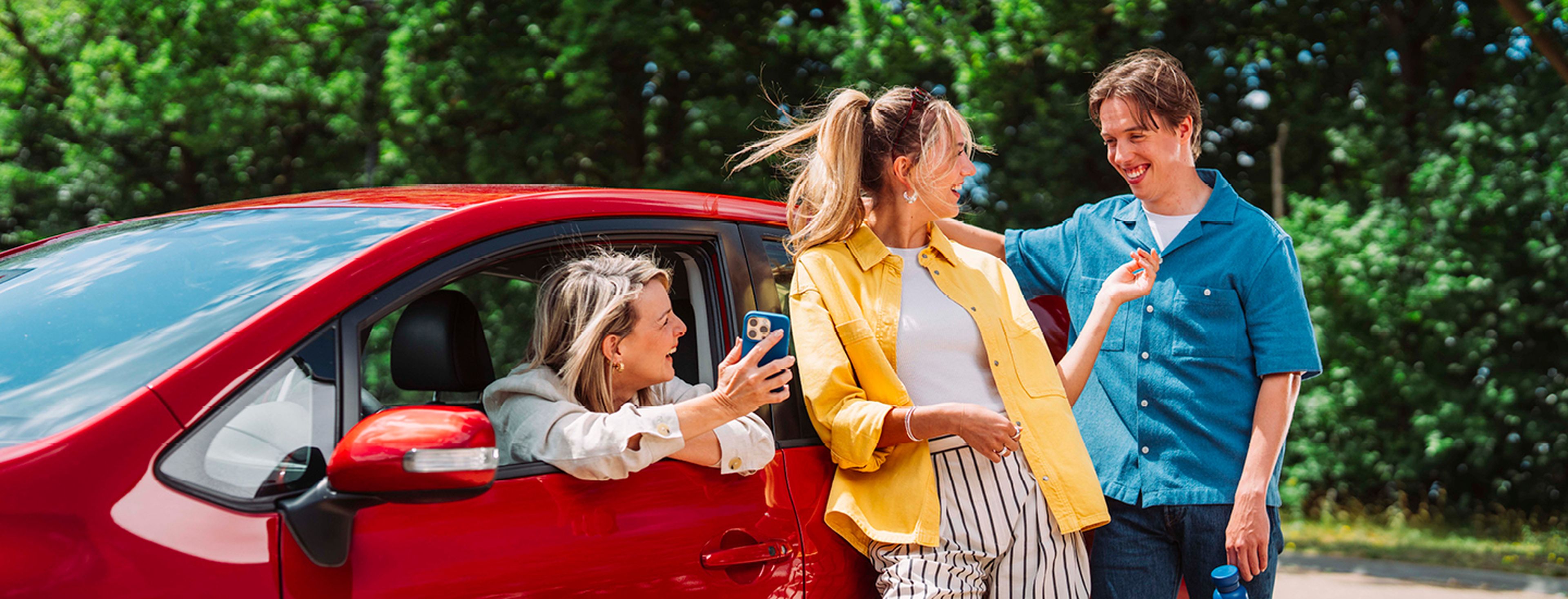 Vrouw zit in de auto en toont haar smartphone aan haar vrienden die tegen de auto leunen